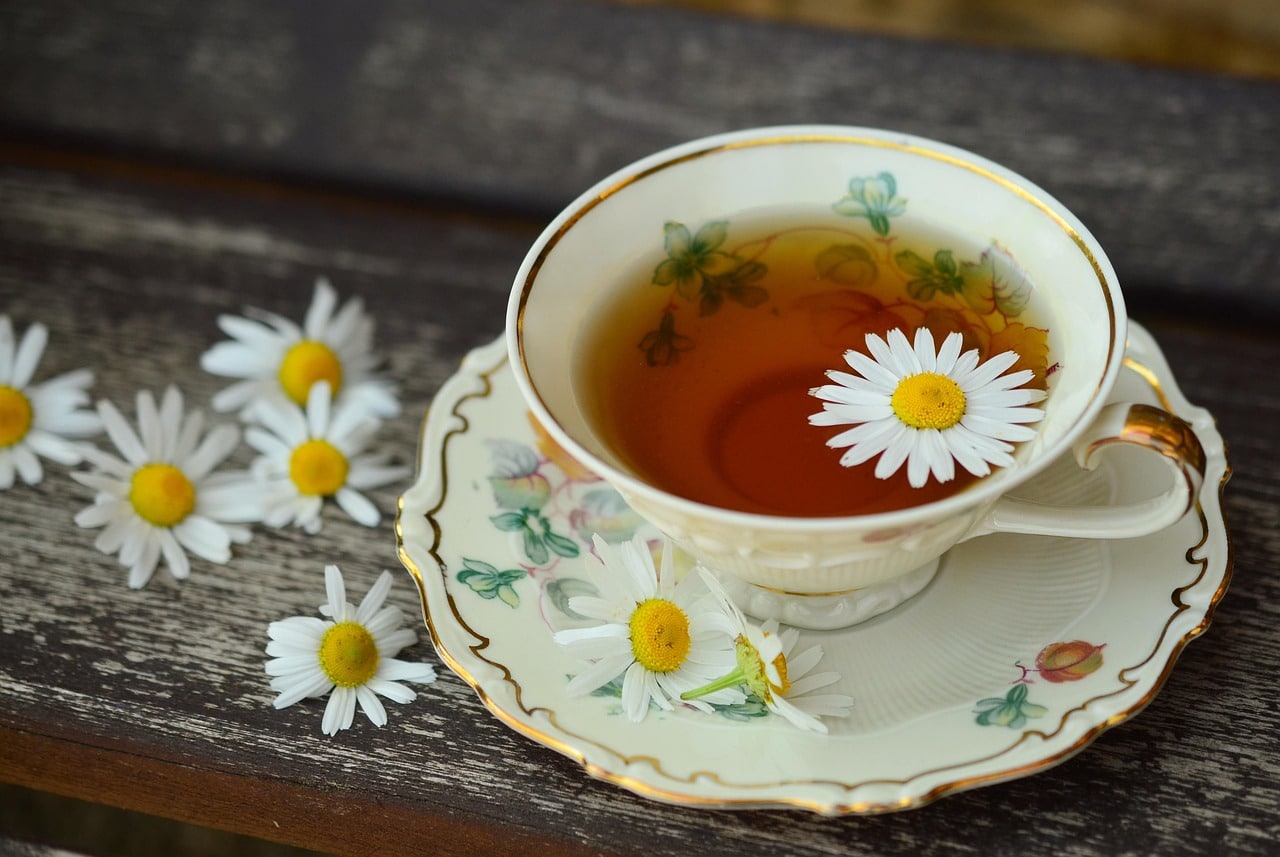 A classic porcelain teacup, likely from the United Kingdom, filled with herbal tea sits on a matching saucer, with a single daisy flower floating in the cup and several others scattered on a wooden surface nearby.
