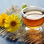 A warm cup of herbal tea in a glass mug, accompanied by two bright yellow calendula flowers, resting on a rustic burlap mat.