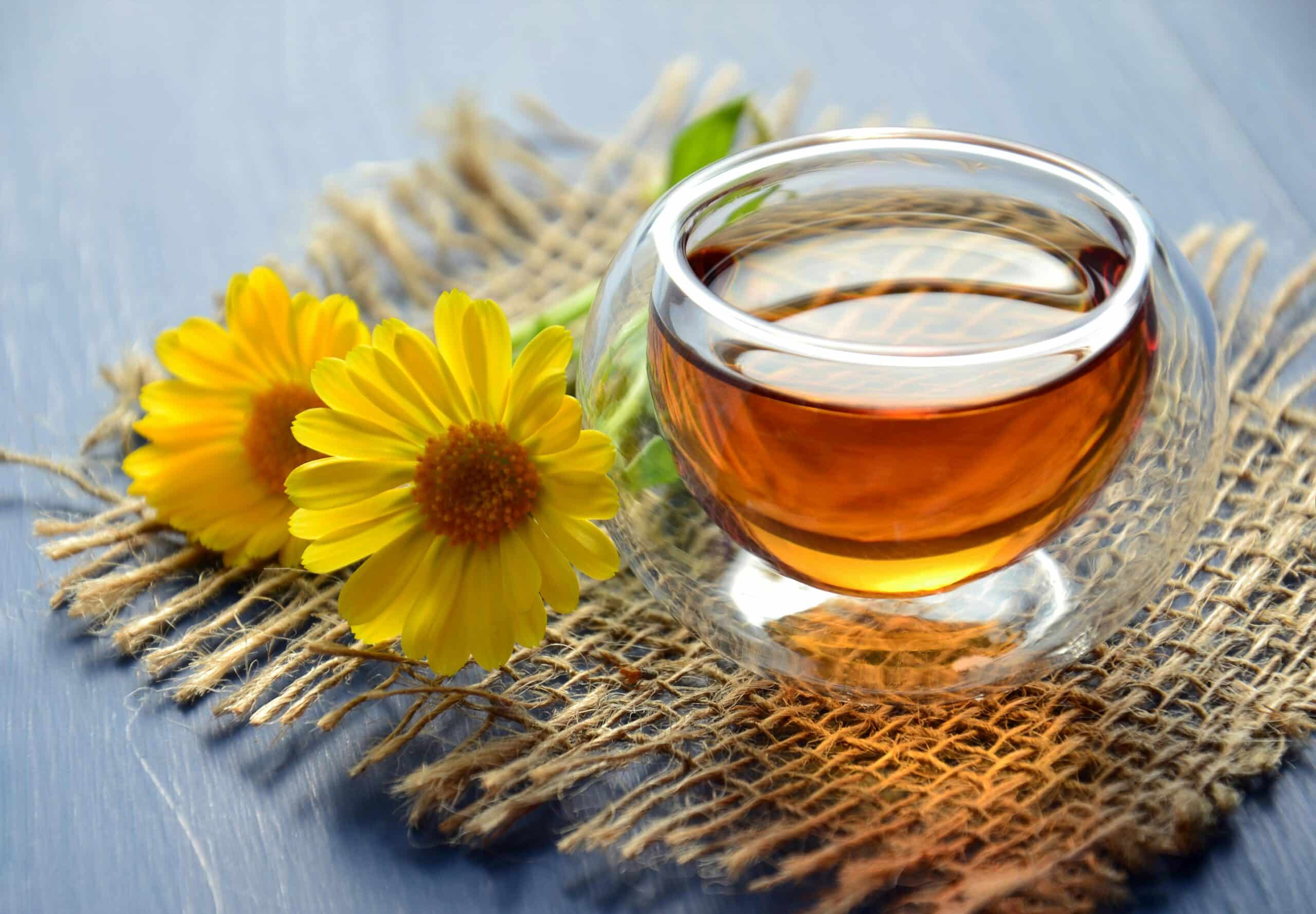A warm cup of herbal tea in a glass mug, accompanied by two bright yellow calendula flowers, resting on a rustic burlap mat.