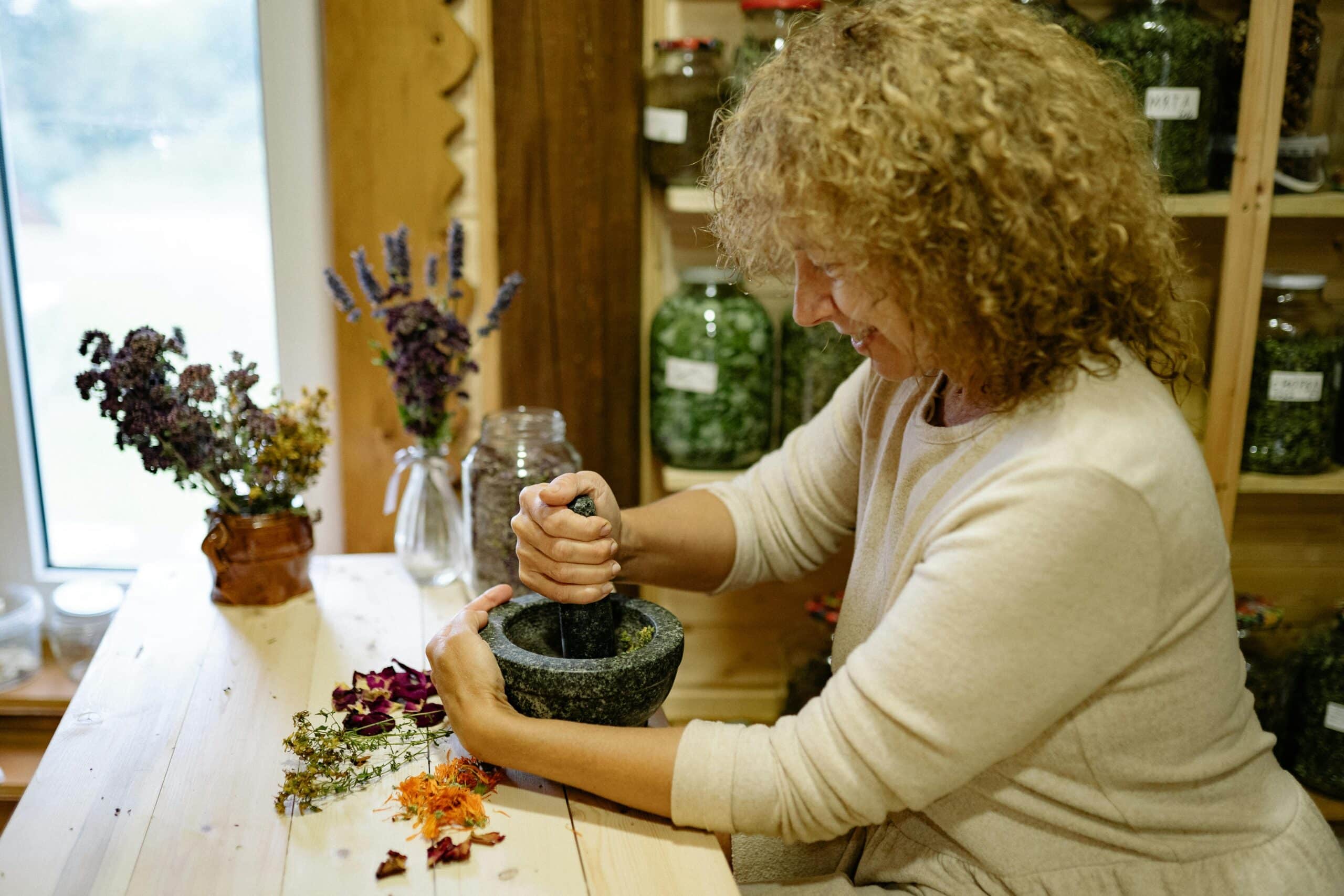 A woman with curly hair grinds dried herbal ingredients in a mortar and pestle, surrounded by various herbs and jars in what appears to be a traditional herbal medicine workshop in the United Kingdom.