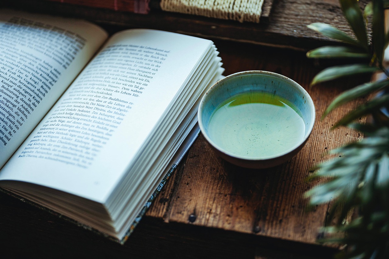 A serene, high-angle shot of an open book resting on a rustic wooden table. To the right of the book sits a small, pale green ceramic bowl filled with a clear liquid. The bowl has a blue and brown rim. In the background, there's a hint of a green plant on the right and some woven material on the left. The scene evokes a sense of quiet relaxation and calm.