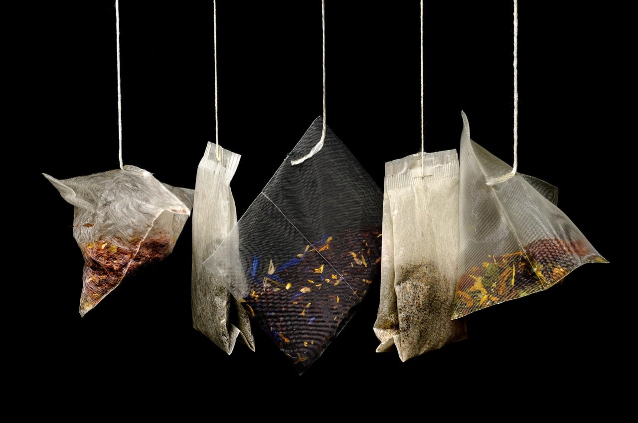 A set of five assorted herbal tea bags hangs suspended against a black background, with the dry herbs and flower petals visible through their mesh and paper filters.