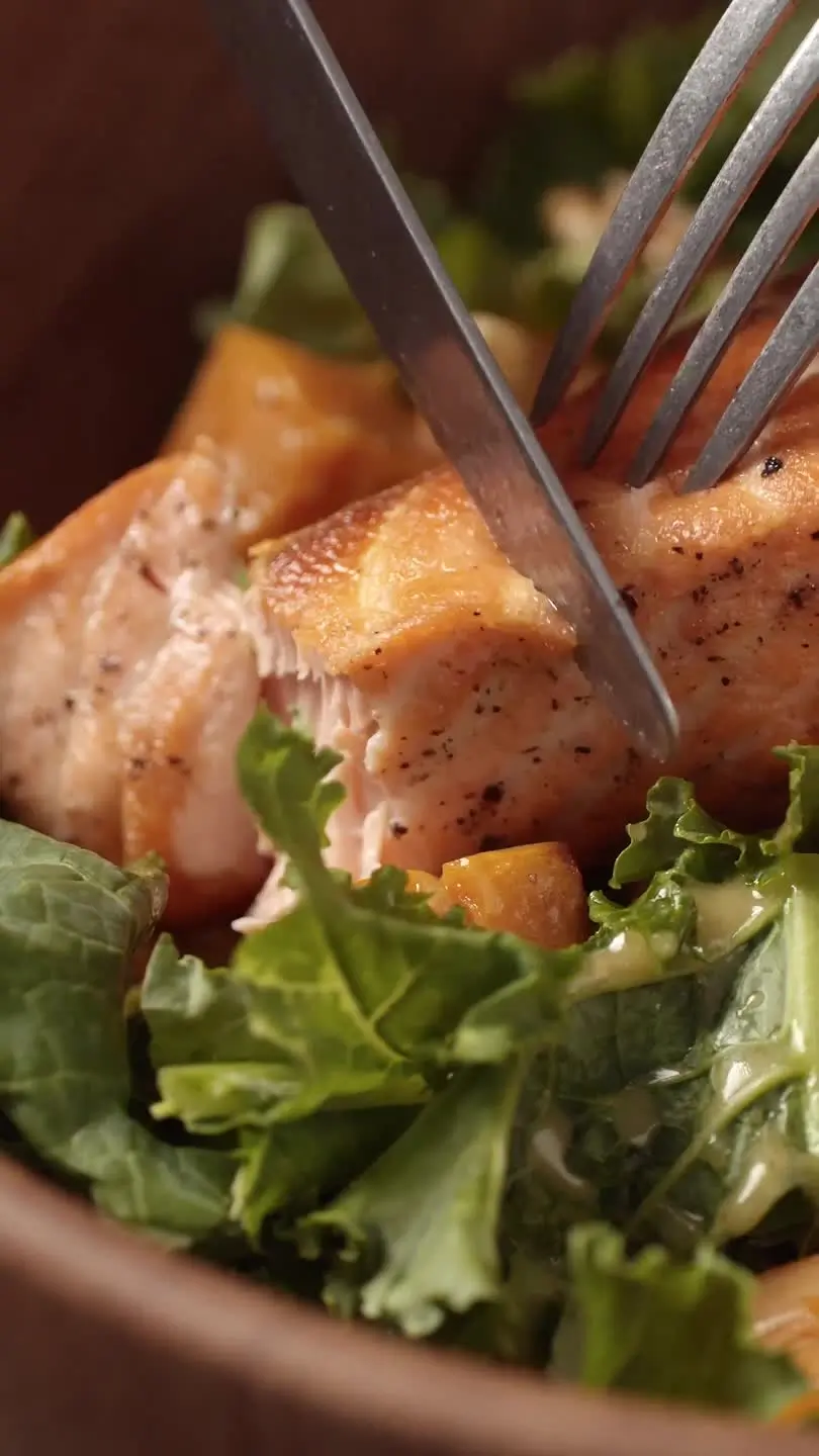 A close-up shows a knife and fork cutting into a perfectly cooked, golden-brown salmon fillet, revealing its flaky, pink interior. The salmon, speckled with black pepper, rests on a bed of green leafy vegetables, possibly kale or lettuce, with a creamy dressing and visible chunks of orange-colored food, suggesting a healthy salmon salad or bowl.