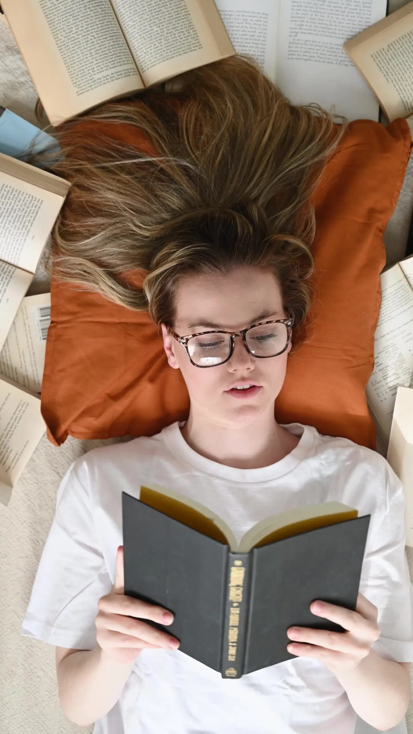 A young woman with blonde hair and glasses lies on her back on a light-colored surface, resting her head on an orange pillow. Her long hair is fanned out above her head, blending with the other open books scattered around her. She is wearing a white t-shirt and holds a dark-covered book open in her hands, engrossed in reading, with her eyes cast down at the pages.