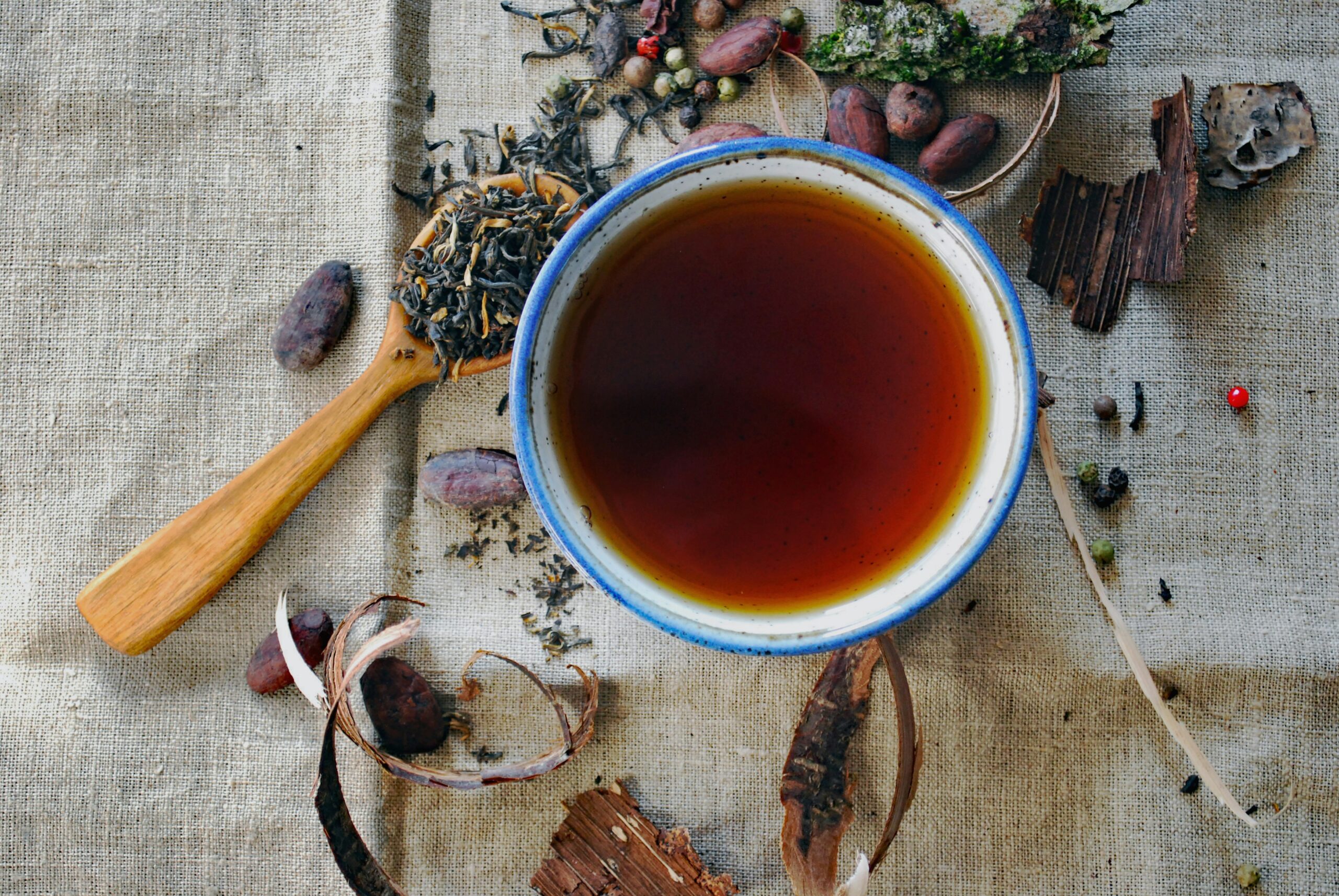 A high-angle shot shows a bowl of dark liquid, likely tea, on a textured, light-colored cloth. To the left, a wooden spoon holds loose, dark tea leaves. Around the bowl are various natural ingredients, including dried pods, bark, and berries.